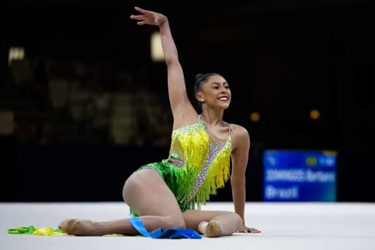 Ginasta Bárbara Domingos está sentada com collant nas cores do Brasil com o braço direito levantado