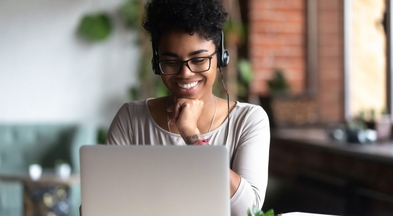 Black woman smart student girl sitting at table in university cafe alone wearing glasses looking at computer screen using headphones listening online lecture improve language skills having good mood
