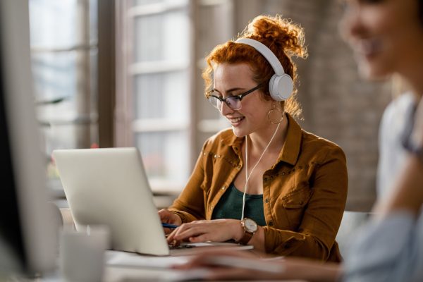 Smiling creative businesswoman listening music on headphones while working on a computer in the office.