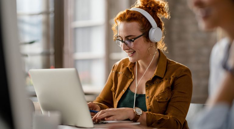 Smiling creative businesswoman listening music on headphones while working on a computer in the office.