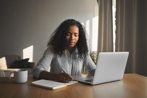 Beautiful mixed race student girl with voluminous hairdo working on home assignment in her room, sitting at wooden desk, using laptop and writing down in copybook. People, technology and education