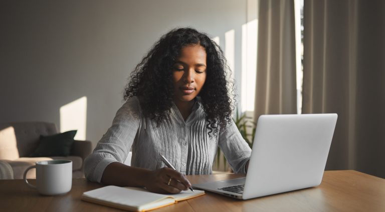 Beautiful mixed race student girl with voluminous hairdo working on home assignment in her room, sitting at wooden desk, using laptop and writing down in copybook. People, technology and education