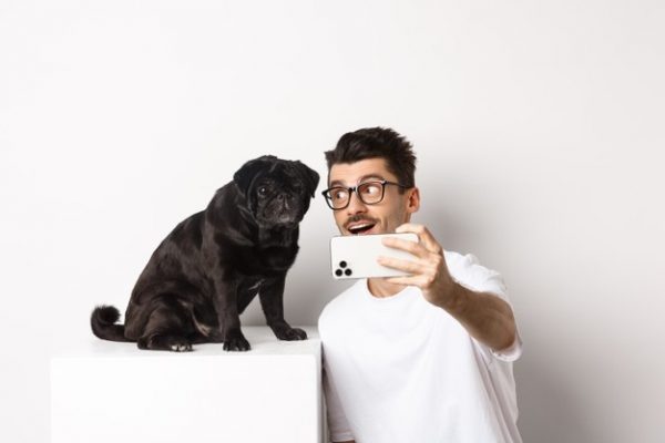 dono-de-cachorro-feliz-tirando-selfie-com-um-lindo-pug-preto-sorrindo-e-olhando-com-amor-para-o-cachorrinho-segurando-um-telefone-celular-branco_1258-32677-600x400