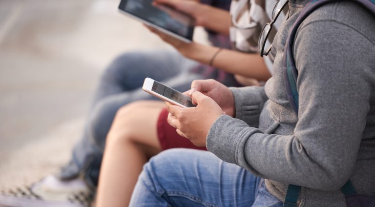 Unrecognizable group of friends in casualwear gathered together outdoors and using their devices, close-up shot