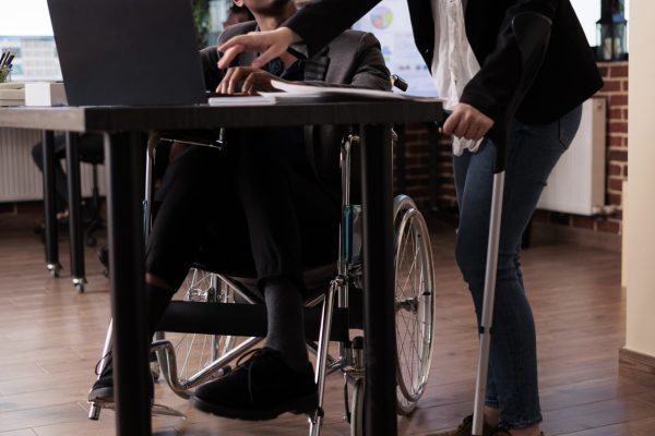 Multiethnic team of businesspeople with chronic impairment working on startup project together. Woman using crutches and man in wheelchair brainstorming ideas in disability friendly office.